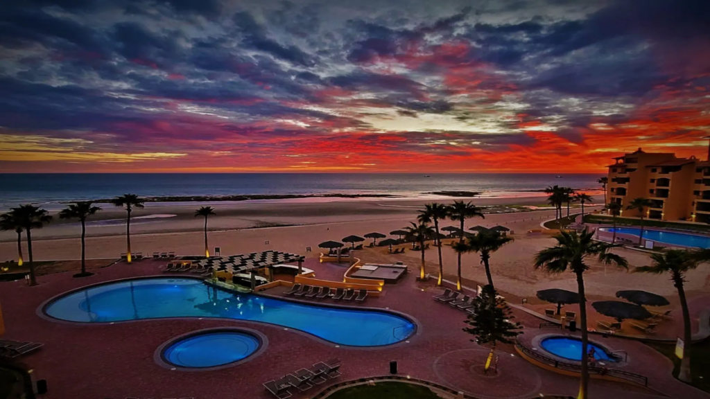 Intense pink and purple sunset clouds over the illuminated resort pool and quiet beach.