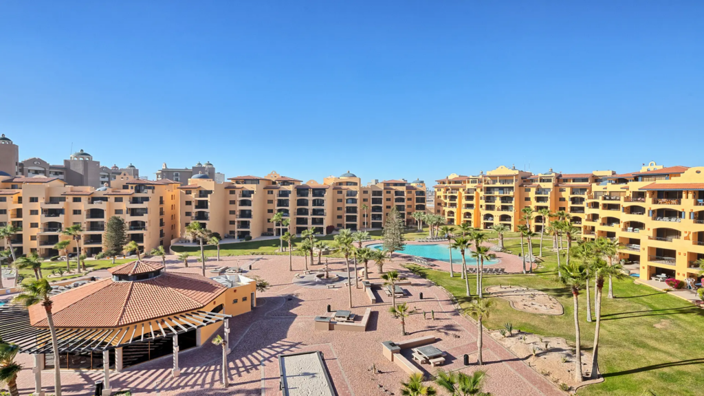 Wide panoramic shot of the golden resort buildings, palm trees, and blue pool under a clear sky.